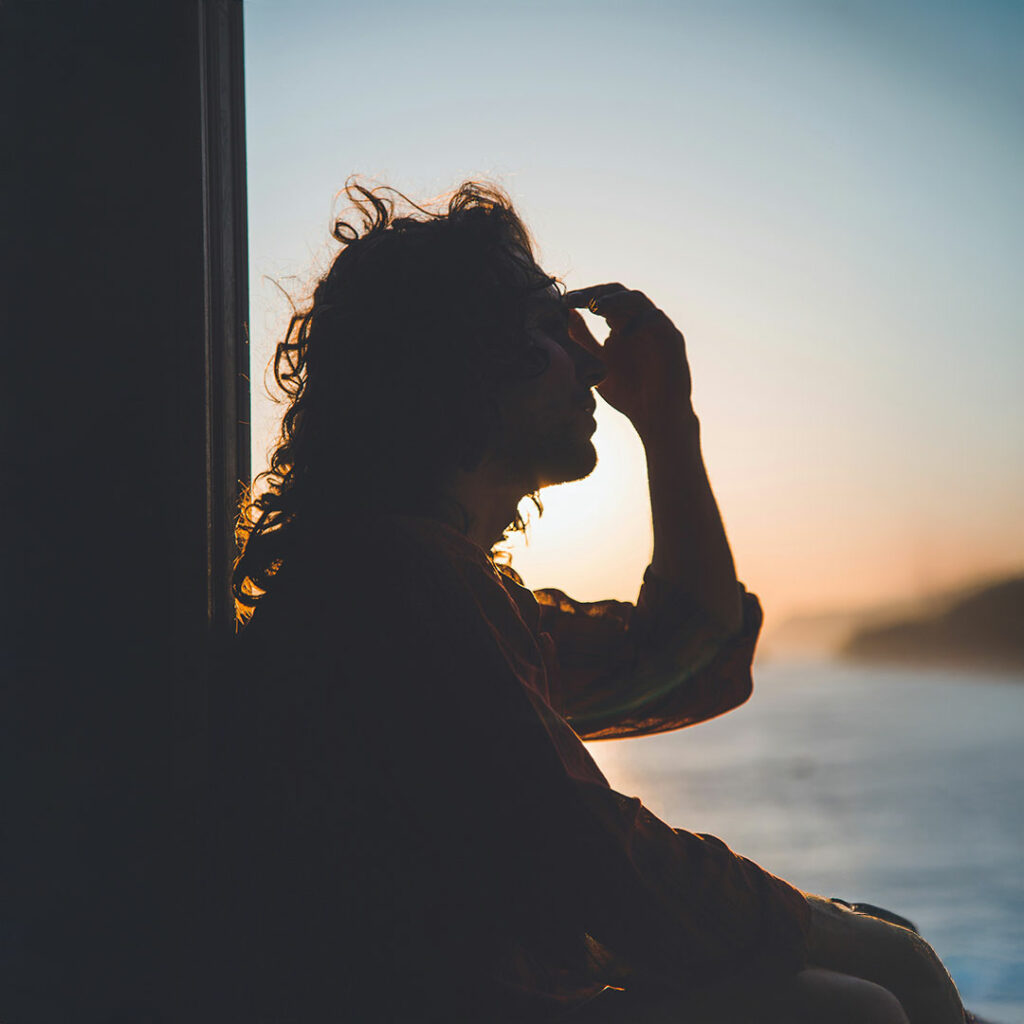 Silhouette of a person touching their forehead while standing near a window at sunset, with a serene ocean and mountain landscape in the background.