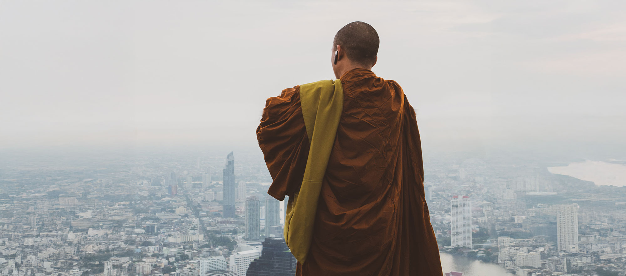 Rear view of a Buddhist monk with wireless earphones, draped in saffron robes, standing above a fog-covered modern city skyline.