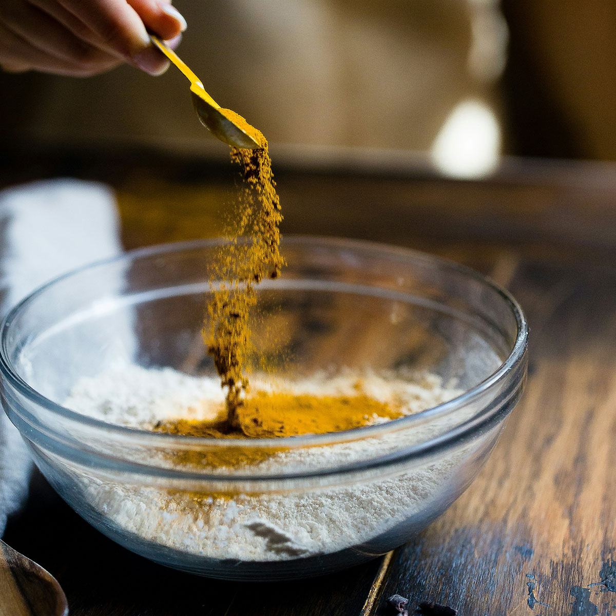 Hand sprinkling turmeric powder into a glass bowl with flour on a wooden kitchen counter.