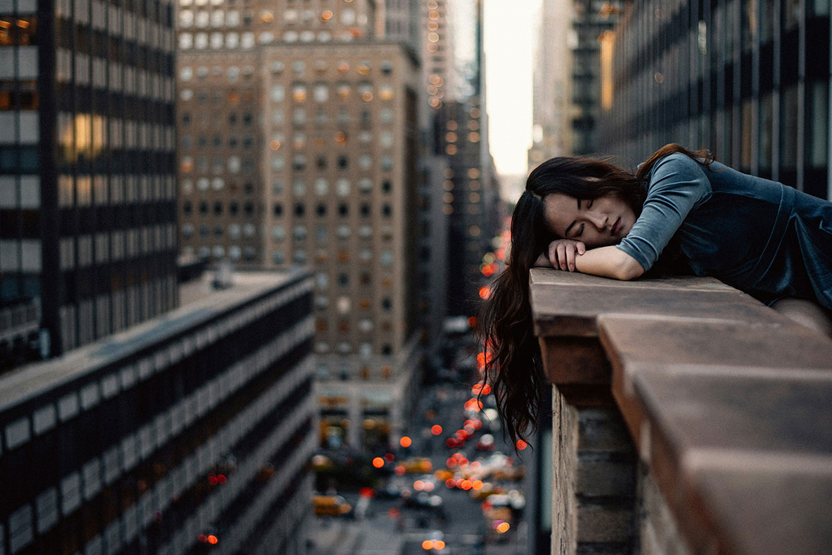 Young woman resting with closed eyes on a rooftop ledge, surrounded by tall buildings and blurred city traffic lights in the background.