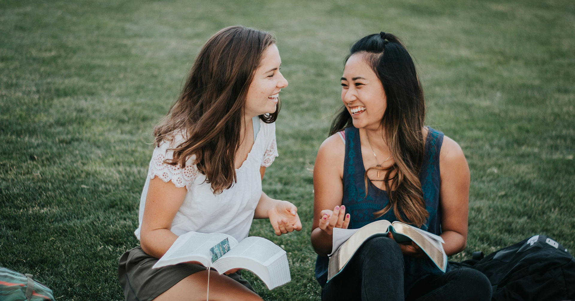 Two young women sitting on grass, smiling and talking while reading books together outdoors.