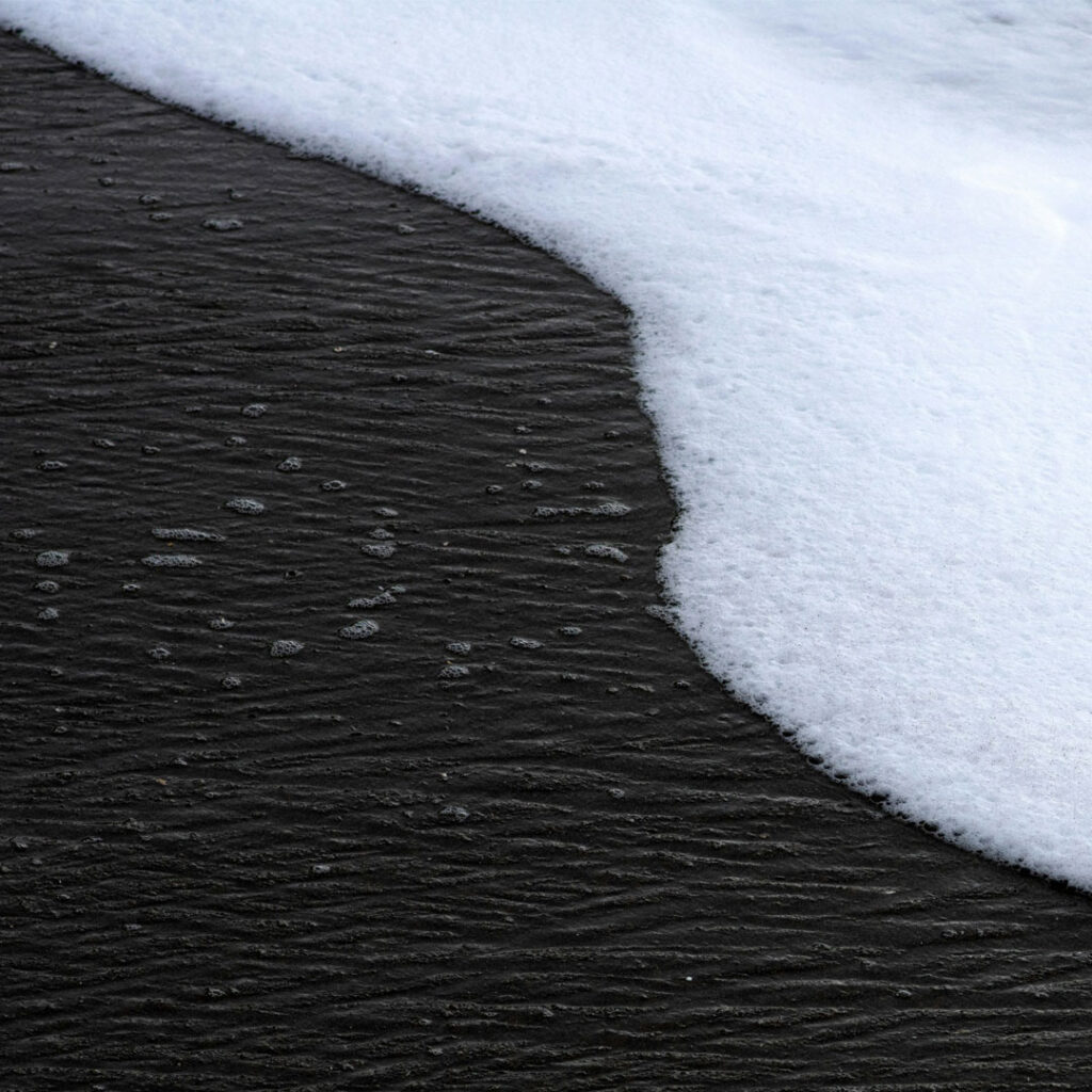 1:1 Close-up of white sea foam on black sand, shot from above, macro photography, detailed photo.