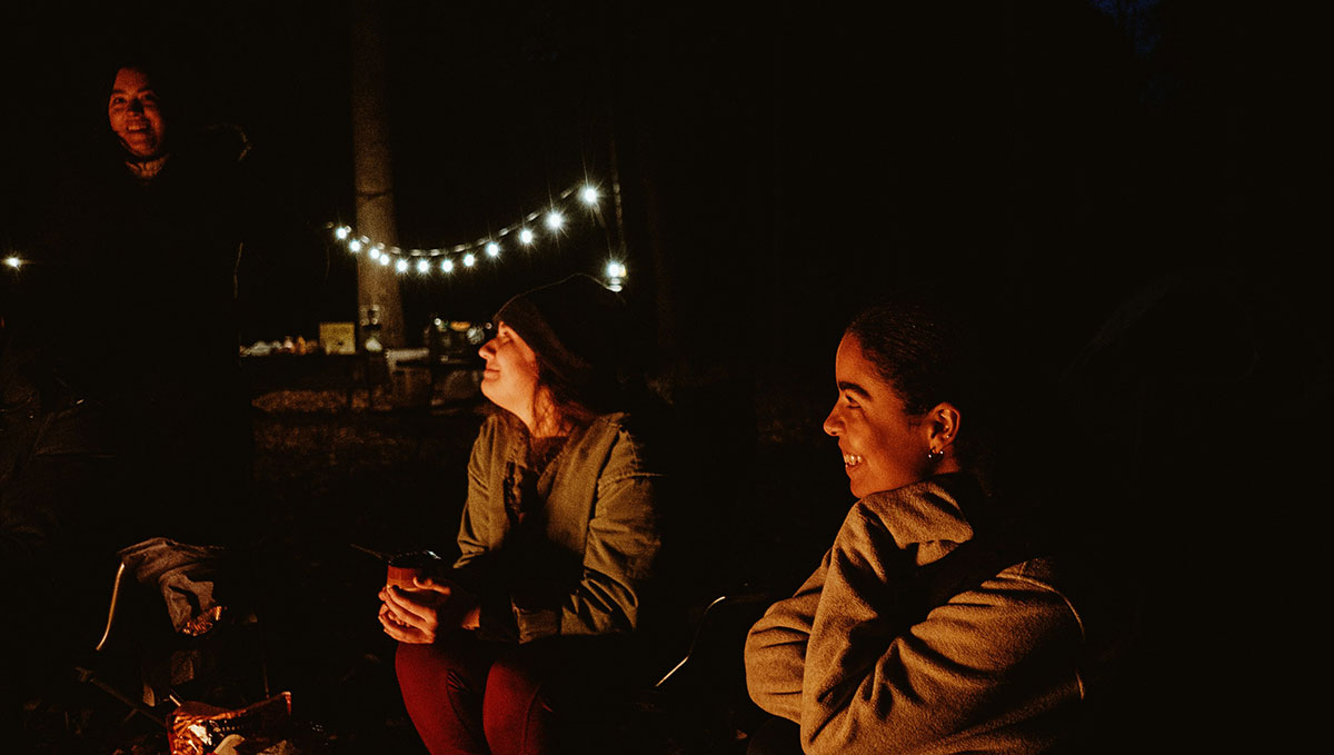 Group of friends sitting around a campfire at night, illuminated by string lights in a forest setting. Keywords (English, comma-separated):