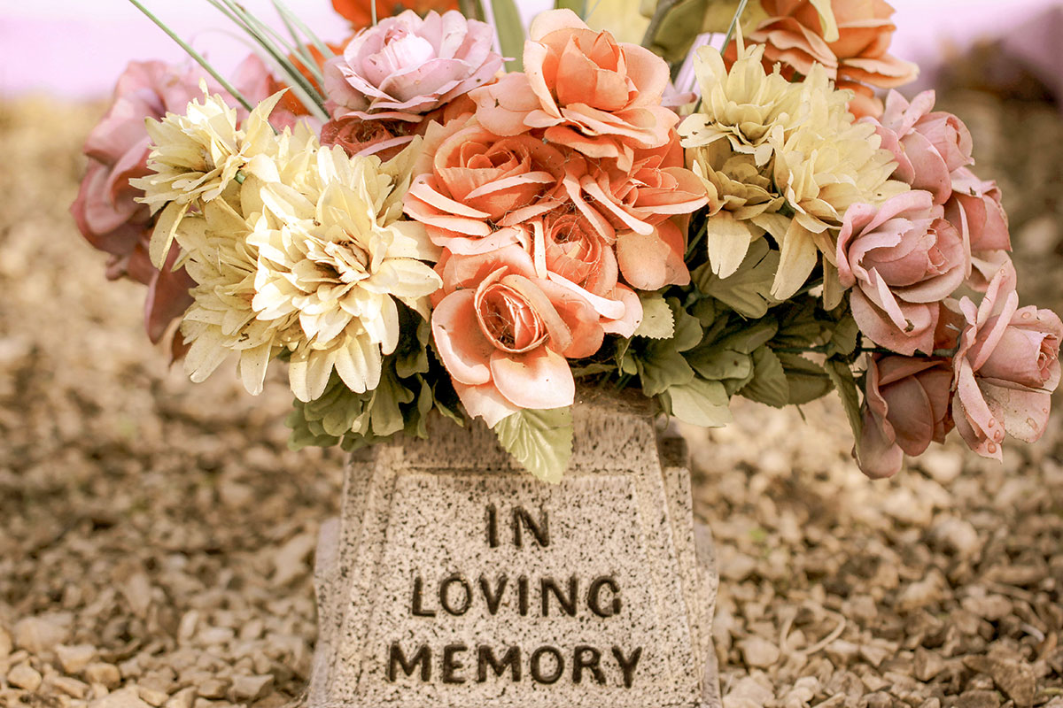 A flower bouquet with pastel-colored roses and dahlias arranged in a stone vase inscribed “In Loving Memory” at a memorial site.