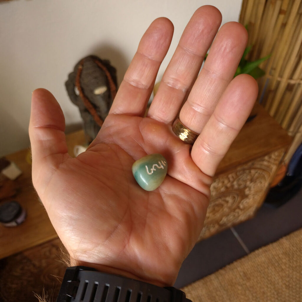 Hand holding a polished green stone with a white handwritten symbol in warm indoor light.